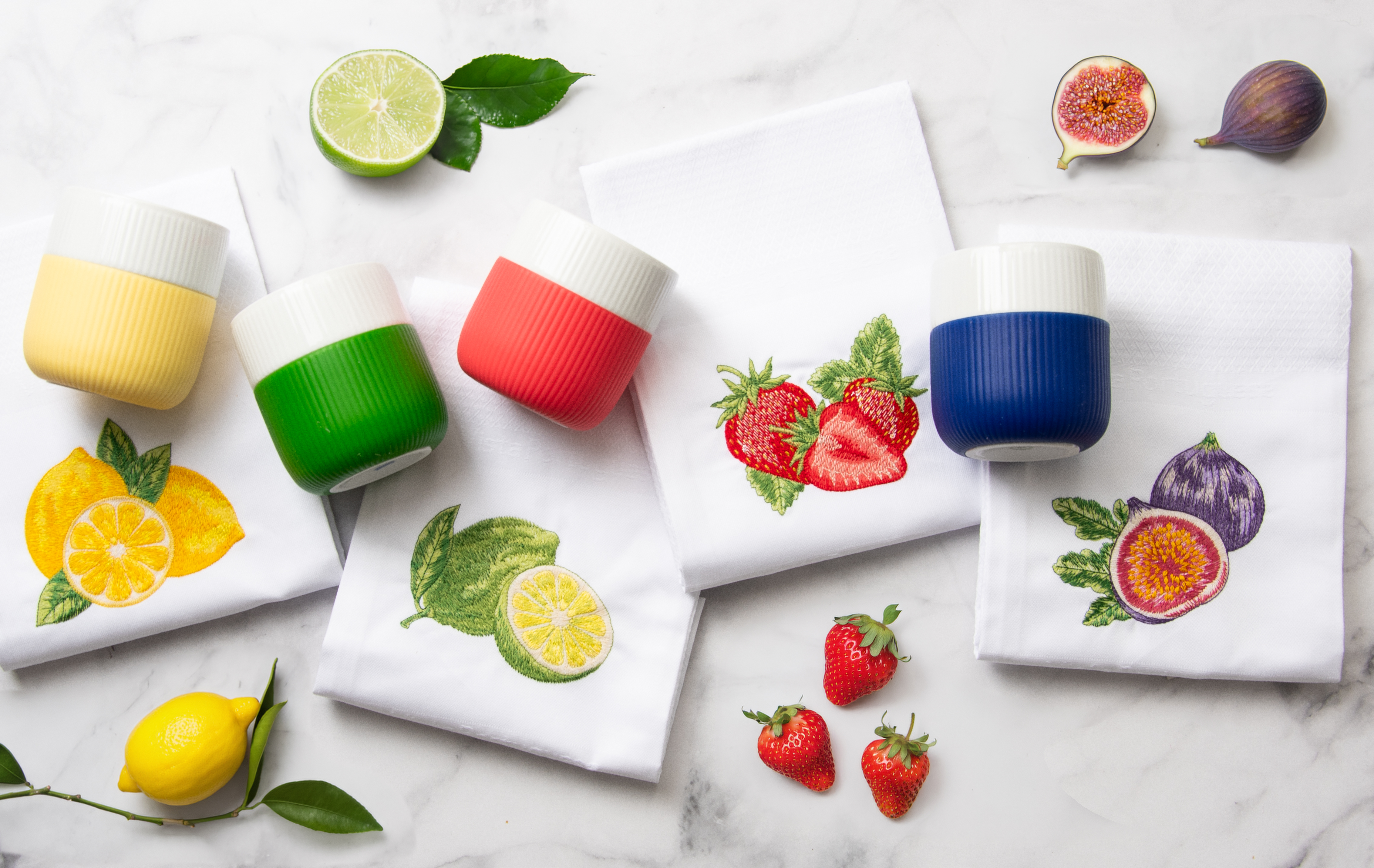 Ein gedeckter Marmortisch von oben fotografiert. Auf dem Tisch liegen die Geschirrtücher Limette, Zitrone, Erdbeeren und Feigen. Dazu liegen jeweils daneben farblich passende Kaffeebecher sowie die entsprechenden Früchte.